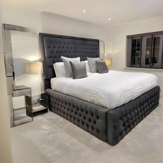 A bedroom with a large dark gray tufted Ivan Hilton bed, white bedding, and modern mirrored nightstands.